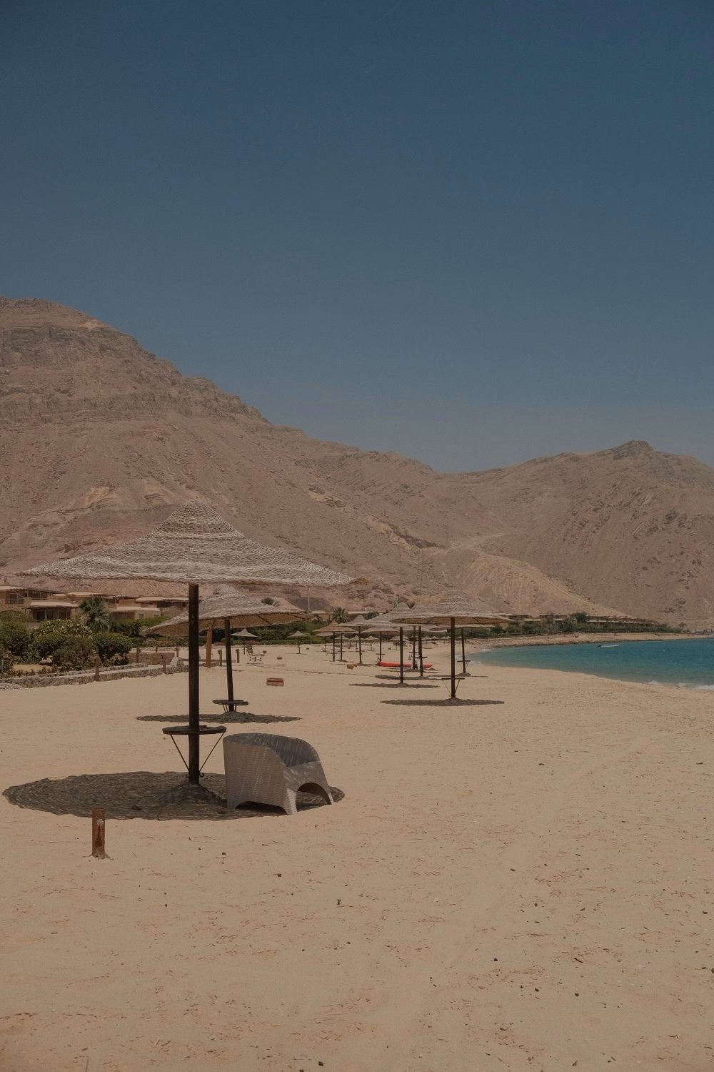 Verlassener Strand mit Fitnessgeräten, türkisem Wasser und kahlen Felsenbergen unter klarem Himmel.