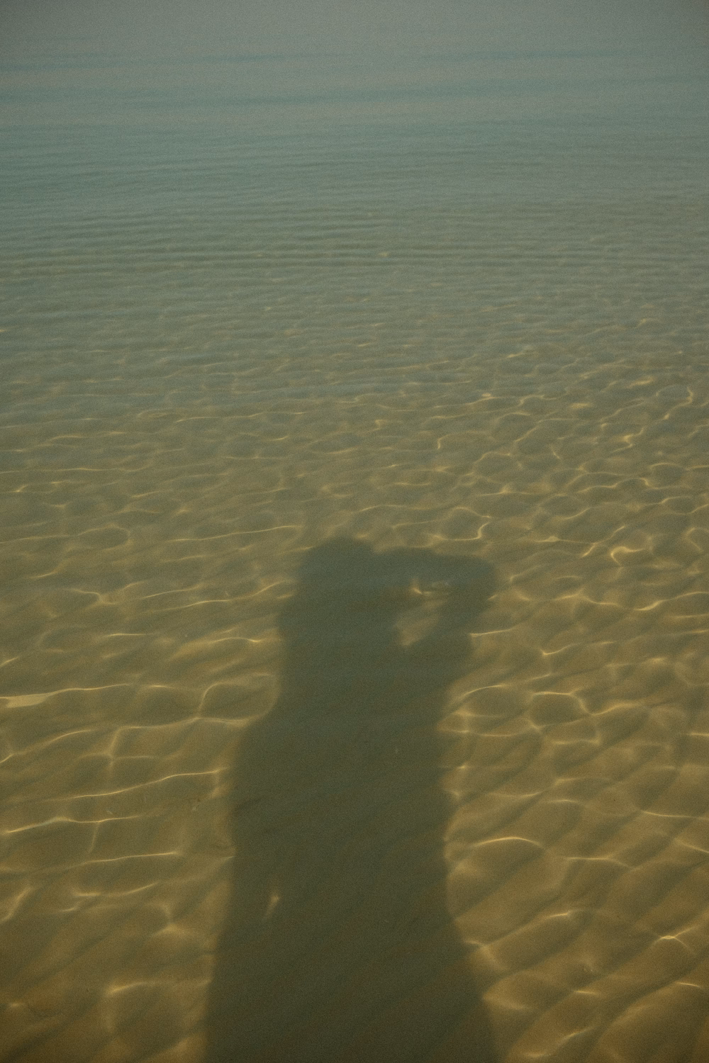 Schatten einer Person auf sandigem Meeresboden mit welligen Mustern und klarem Wasser.