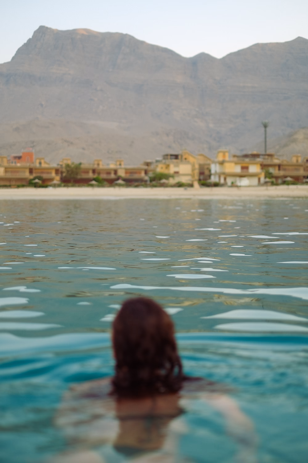 Person im türkisfarbenen Wasser blickt auf gelbe Häuser und Berge in einer Wüstenlandschaft.