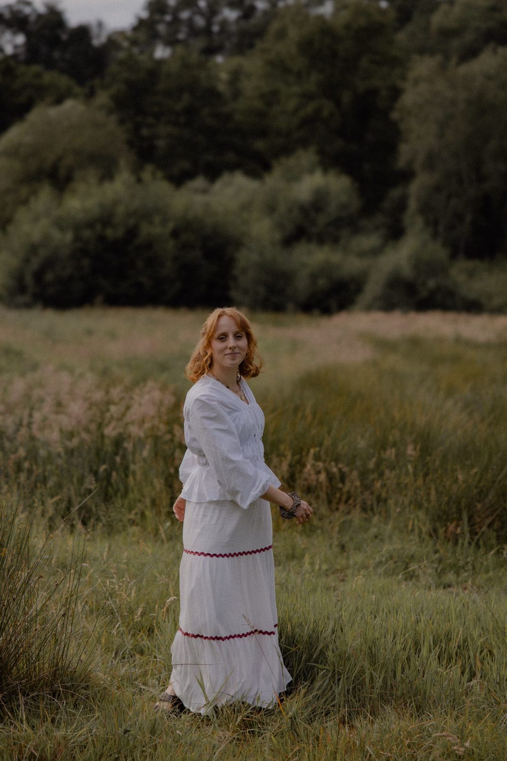 Frau mit rötlichen Haaren in weißem Kleid steht in einem Getreidefeld mit Waldkulisse im Hintergrund.