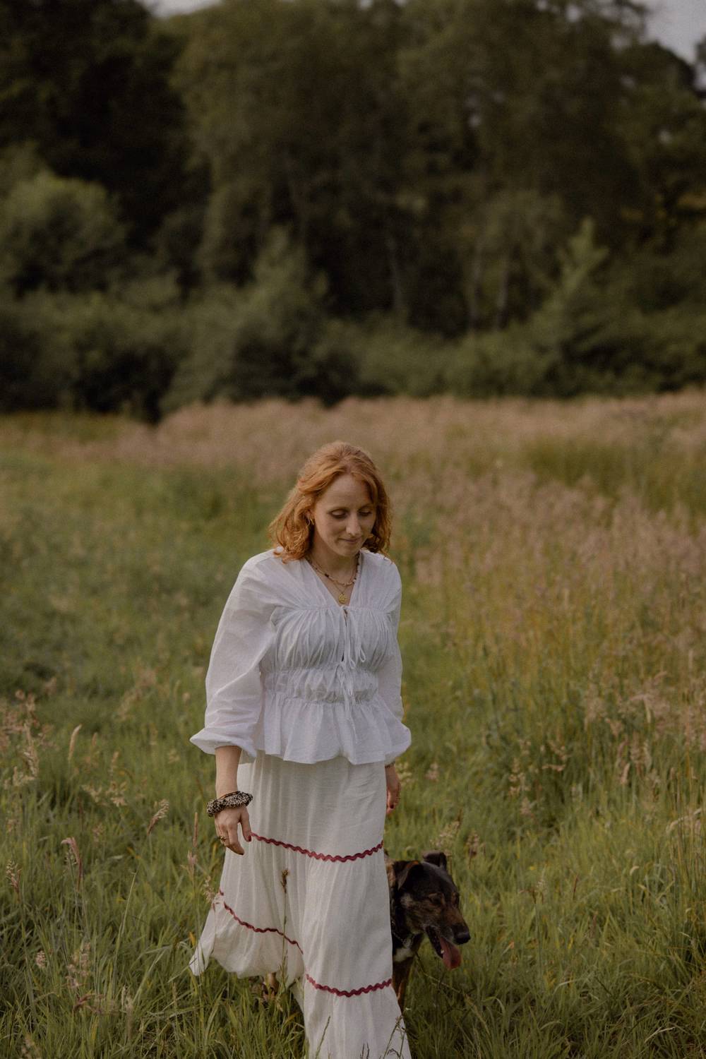 Frau in weißem Kleid mit rötlichem Haar führt einen schwarzen Hund an der Leine durch ein grünes Feld.