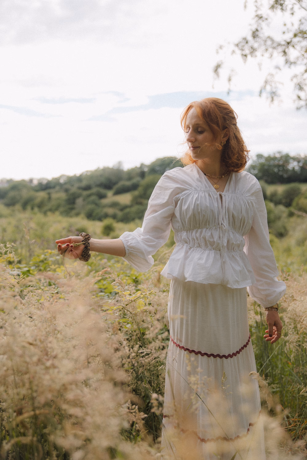Frau in weißem Kleid pflückt Brombeeren in ländlicher Landschaft bei Sonnenschein.