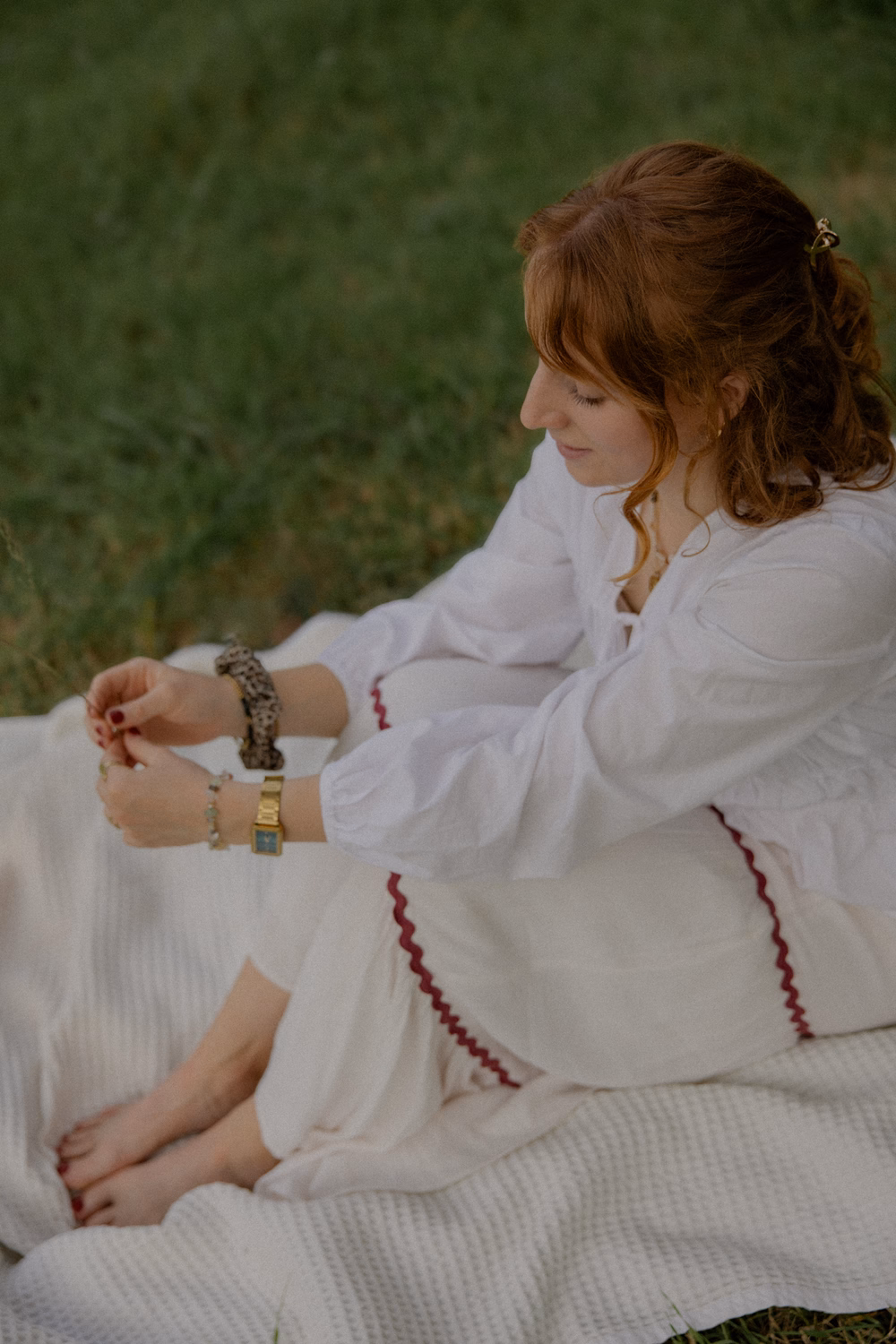 Frau mit rötlichen Haaren sitzt auf weißer Decke und trägt mehrere Armbänder in der Natur.