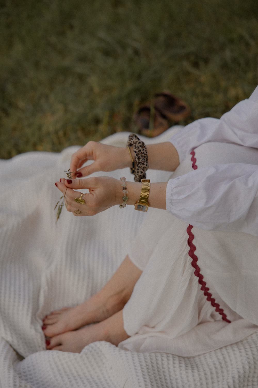 Frau in weißem Kleid mit roten Details, Armbändern und dunklem Nagellack hält Blume auf grünem Hintergrund.