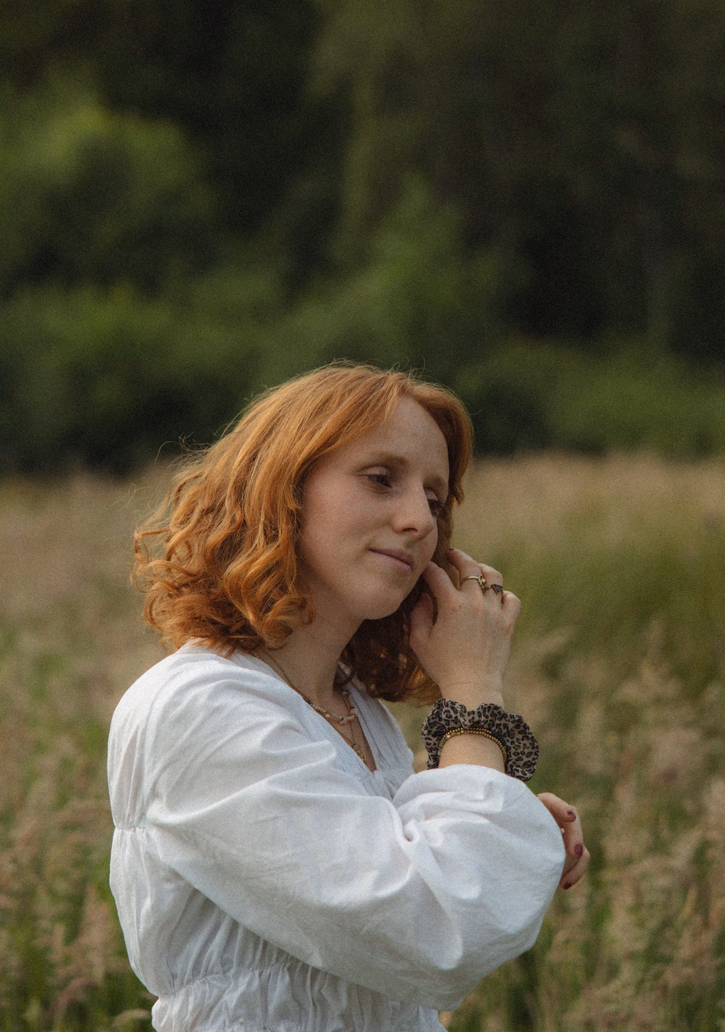Frau mit rötlichem lockigem Haar in weißem Kleid steht nachdenklich in einem Feld mit unscharfem grünem Hintergrund.