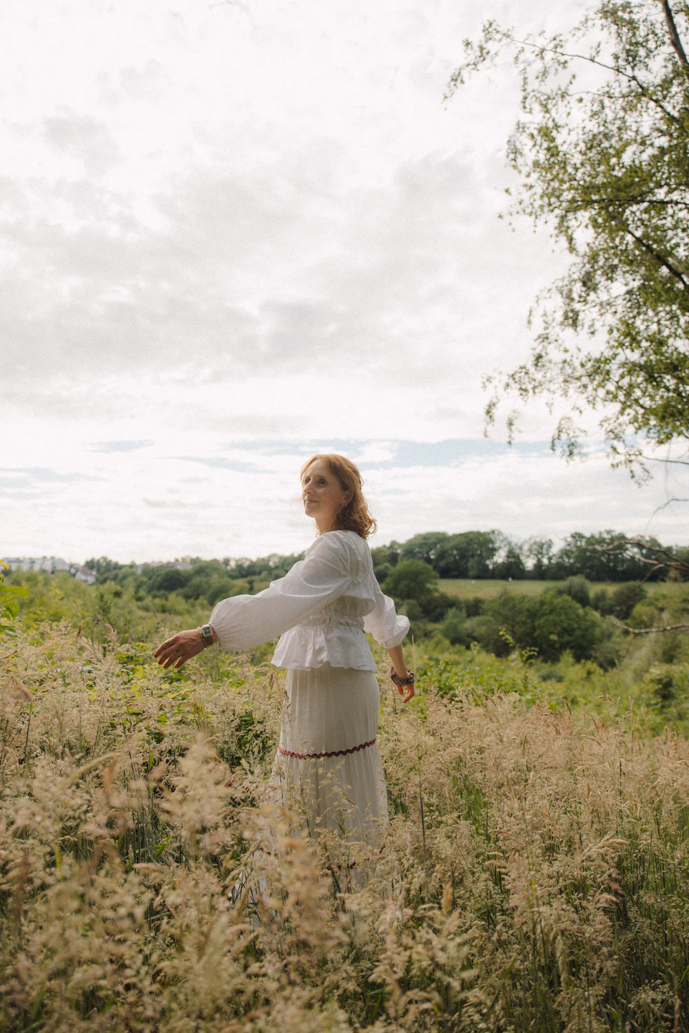 Frau in weißem Kleid steht in einem Feld mit Blick auf eine grüne Landschaft und einen See in der Ferne.